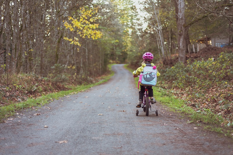 Tips to Transition from Training Wheels a Young Girl Wearing a School Backpack Riding a Bike with Training Wheels Down a Pathway Lined with Trees